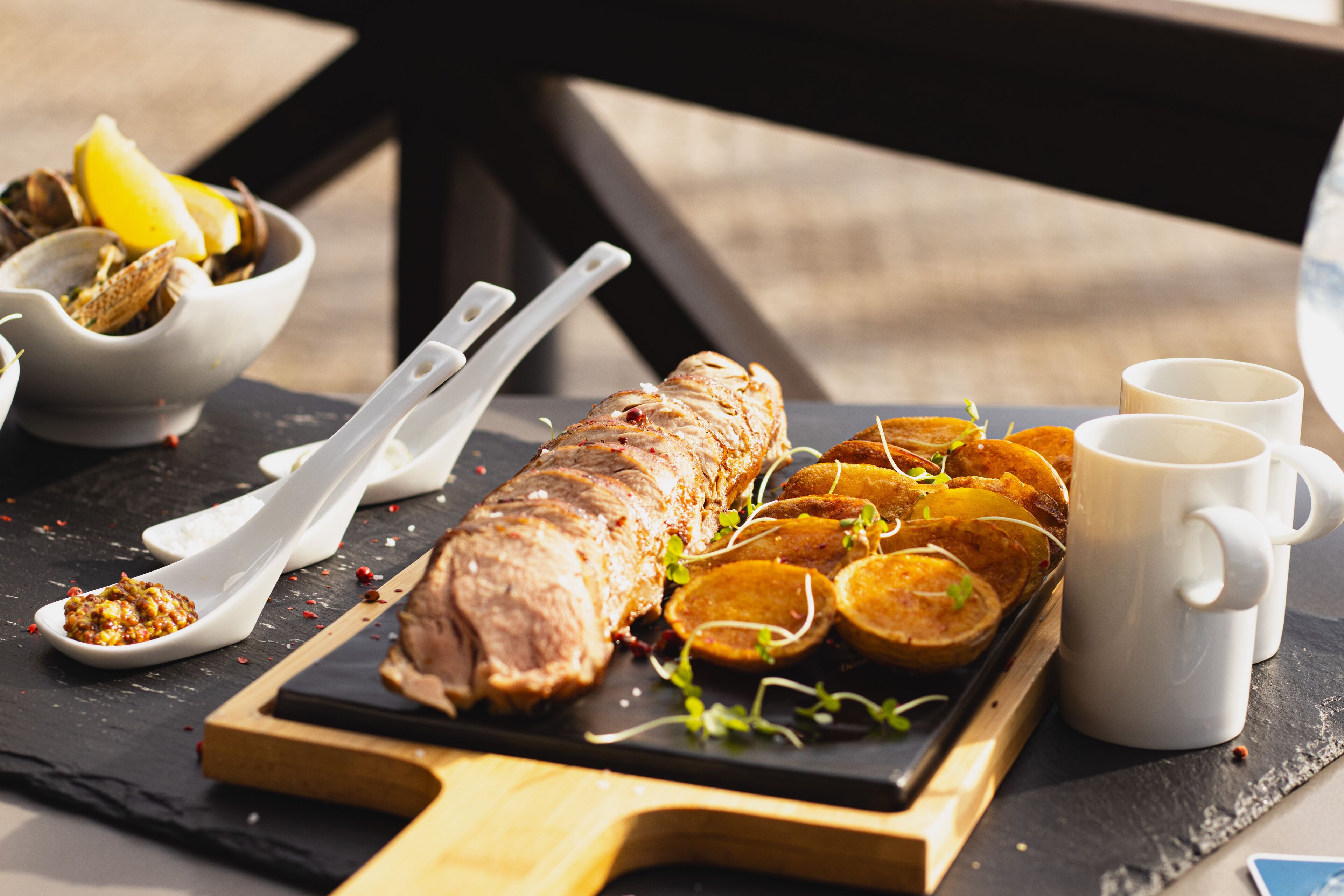 A roasted meat dish on a wooden board, accompanied by sliced vegetables and two white cups on a black slate surface.