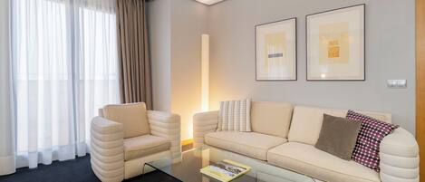Una habitación de hotel moderna con un sofá beige, un sillón a juego y una mesa de centro de vidrio. Hay dos obras de arte enmarcadas en la pared y un ventanal con cortinas traslúcidas.