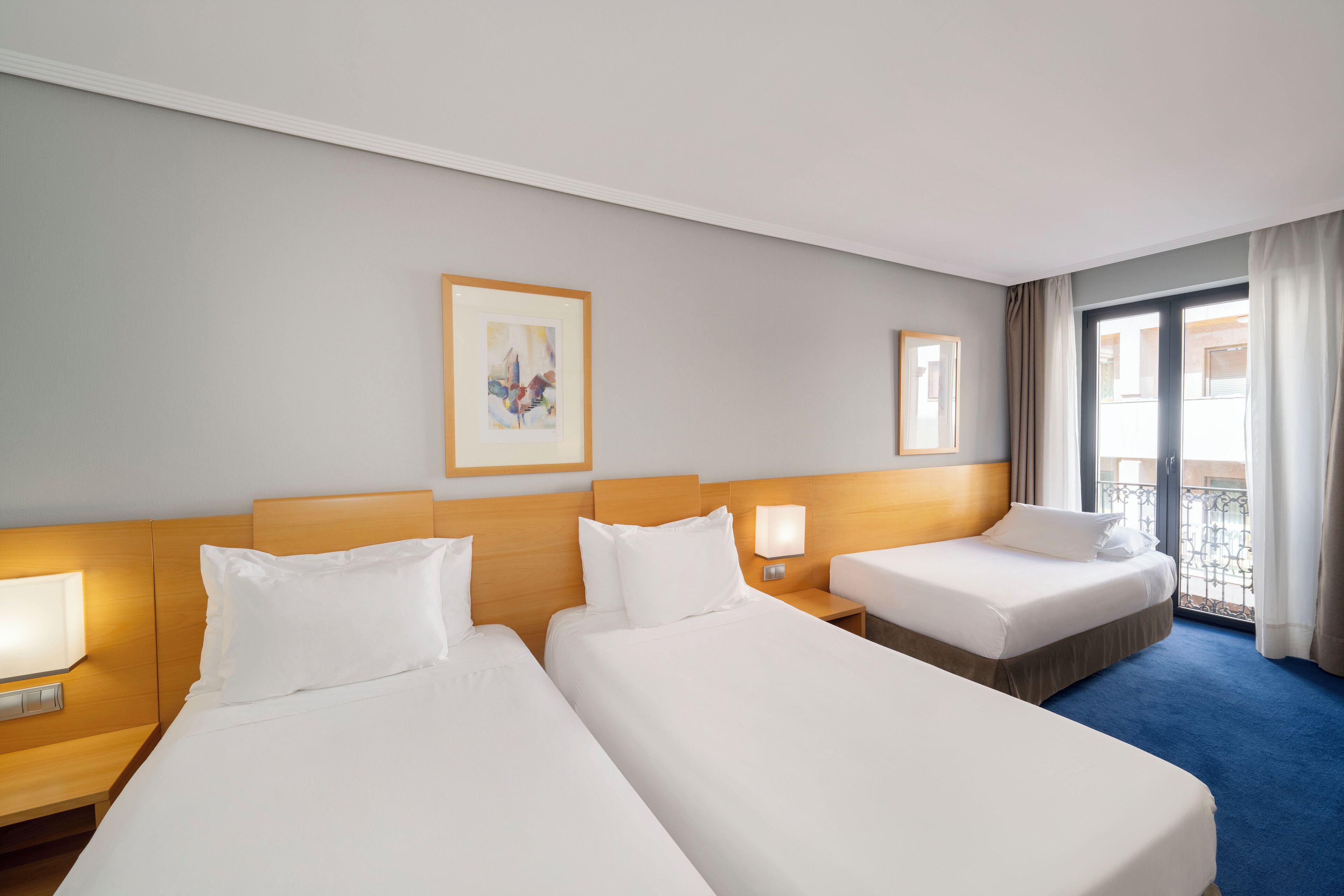 A hotel room with two beds, a nightstand with a lamp, a painting on the wall, and a balcony with curtains.