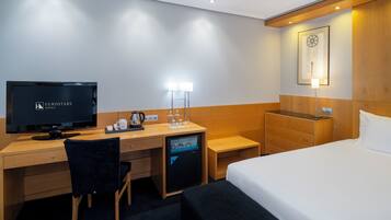 Habitación de hotel con una cama, un escritorio con televisor, una silla, un aparador y una mesita de noche con lámpara.