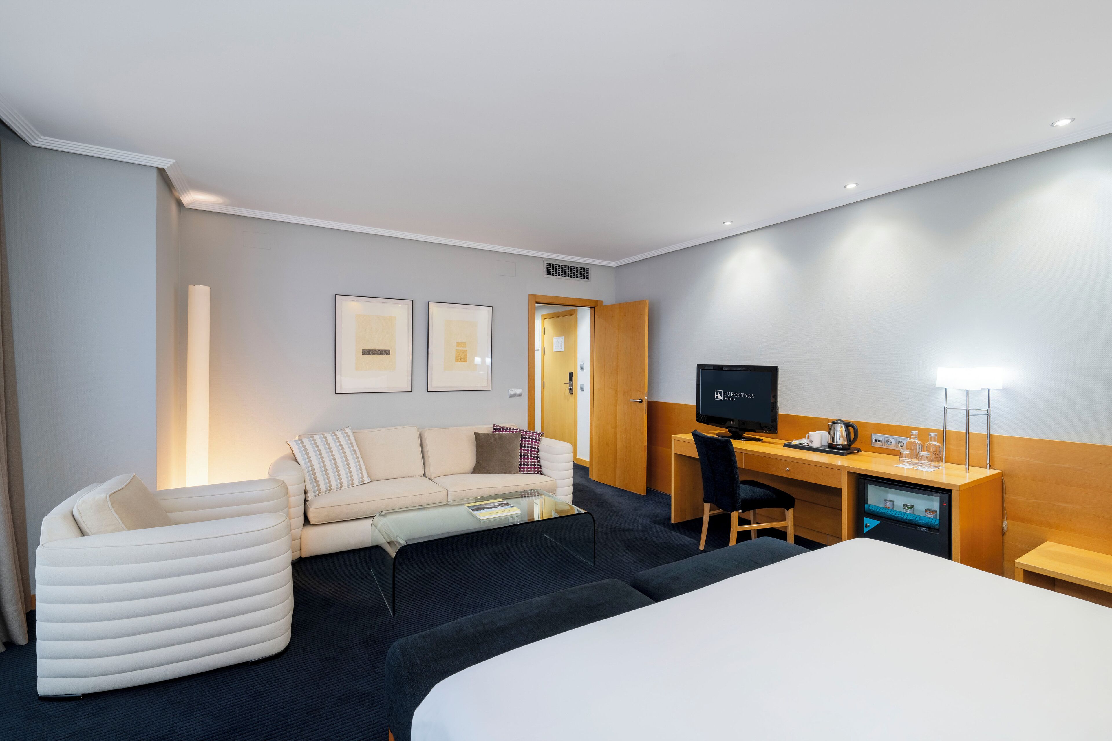 A hotel room with a bed, a sofa, a desk with a computer, a TV, and a minibar.
