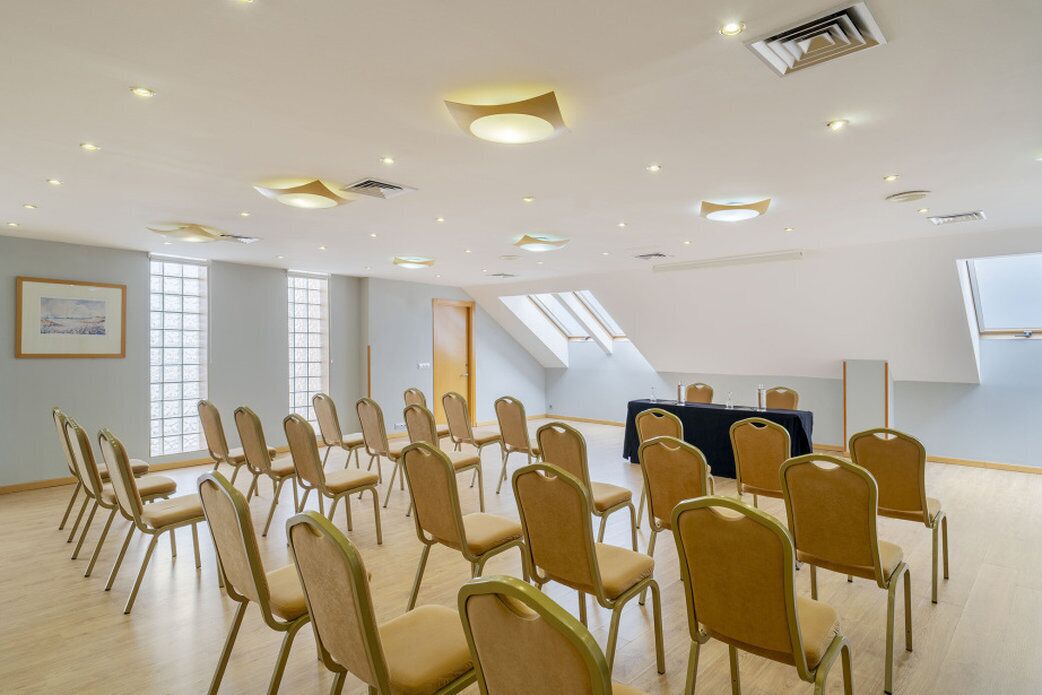 Un salón de conferencias con filas de sillas, una mesa con mantel oscuro y tragaluces.