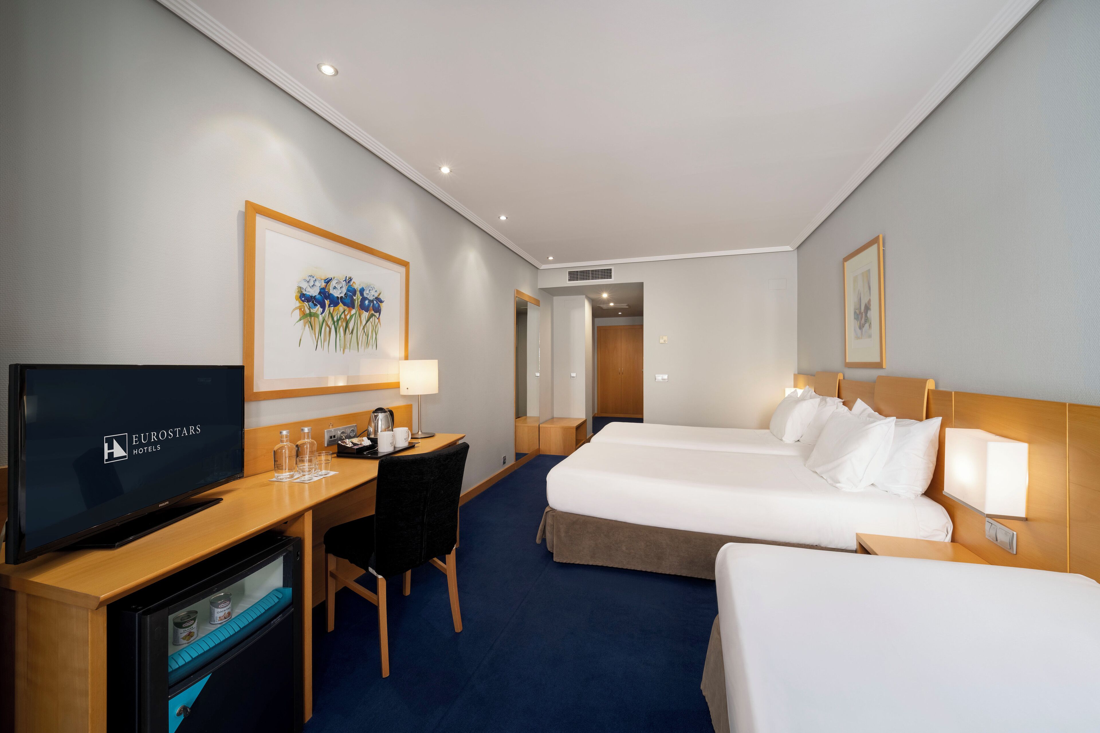 Una habitación de hotel con televisión, escritorio, dos camas y obras de arte en las paredes.