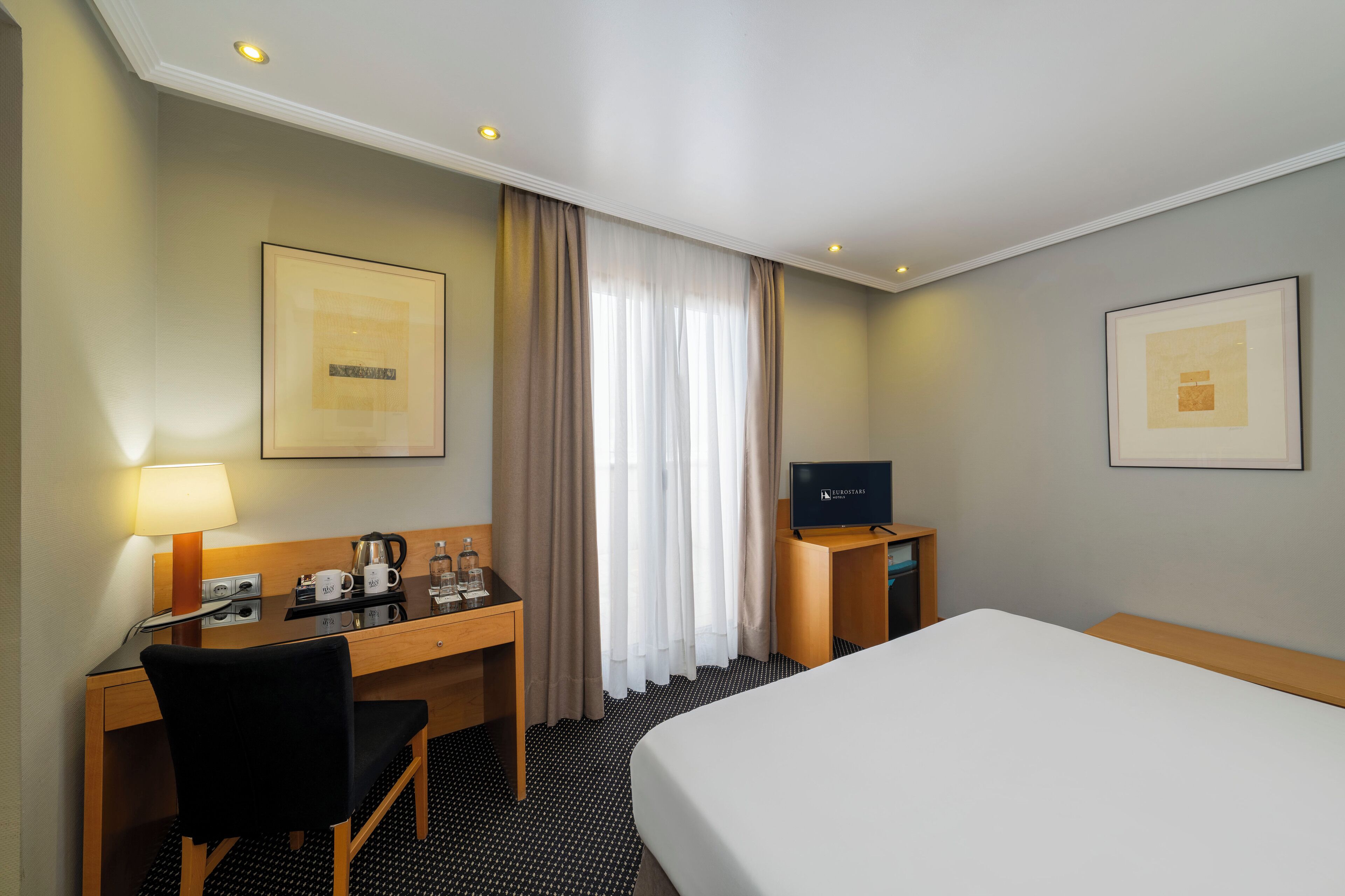A hotel room with a bed, a desk with a chair, a television, a coffee maker, and a lamp.