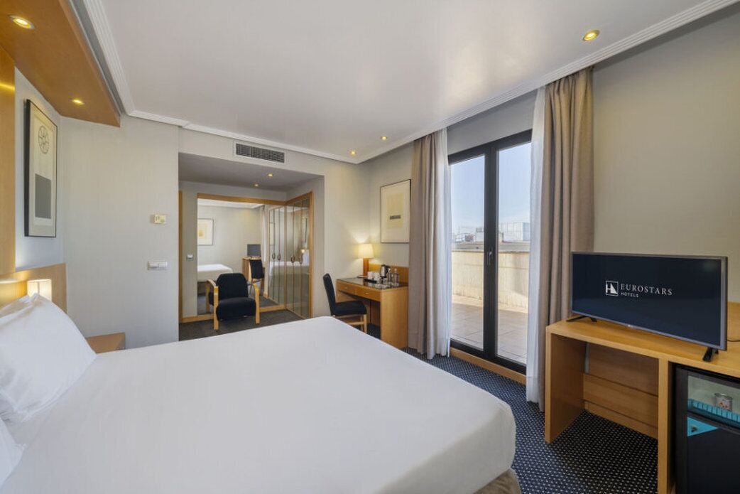 A hotel room with a large bed, a desk with a chair, a television, and a view of the city through the window.