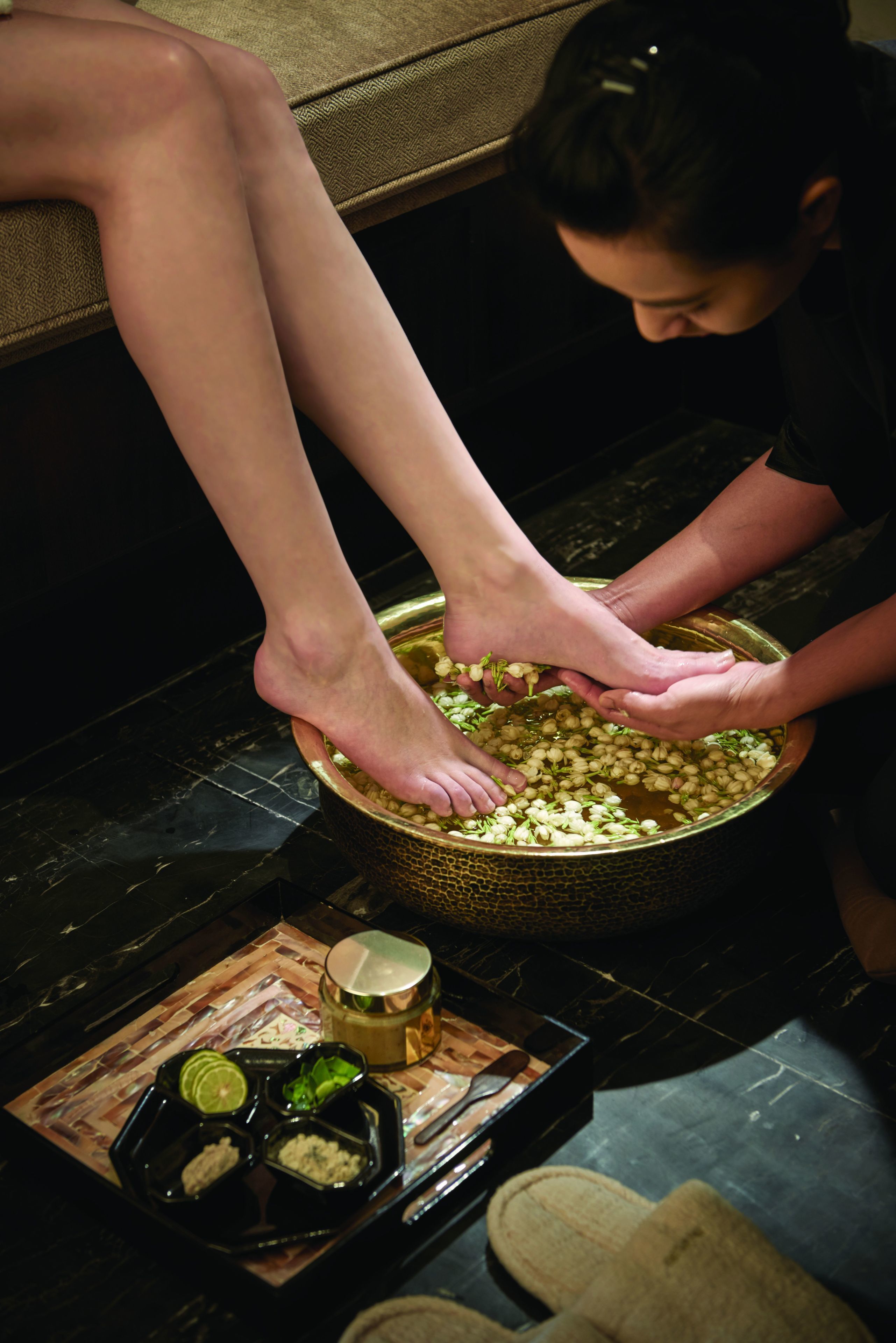 A person soaking their feet in a large bowl filled with water and herbs, with a tray of condiments nearby.