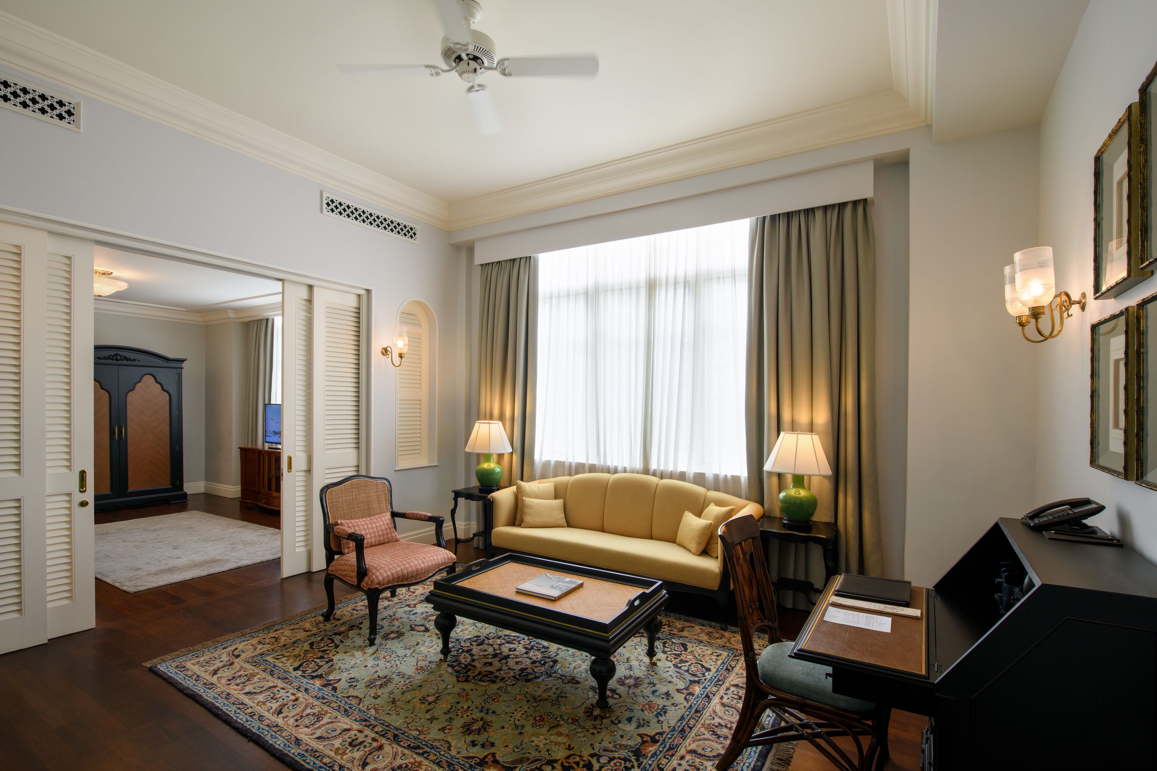 A hotel room with a sofa, armchairs, a coffee table, a desk, and a computer. There is a window with curtains, a ceiling fan, and a wall-mounted lamp.