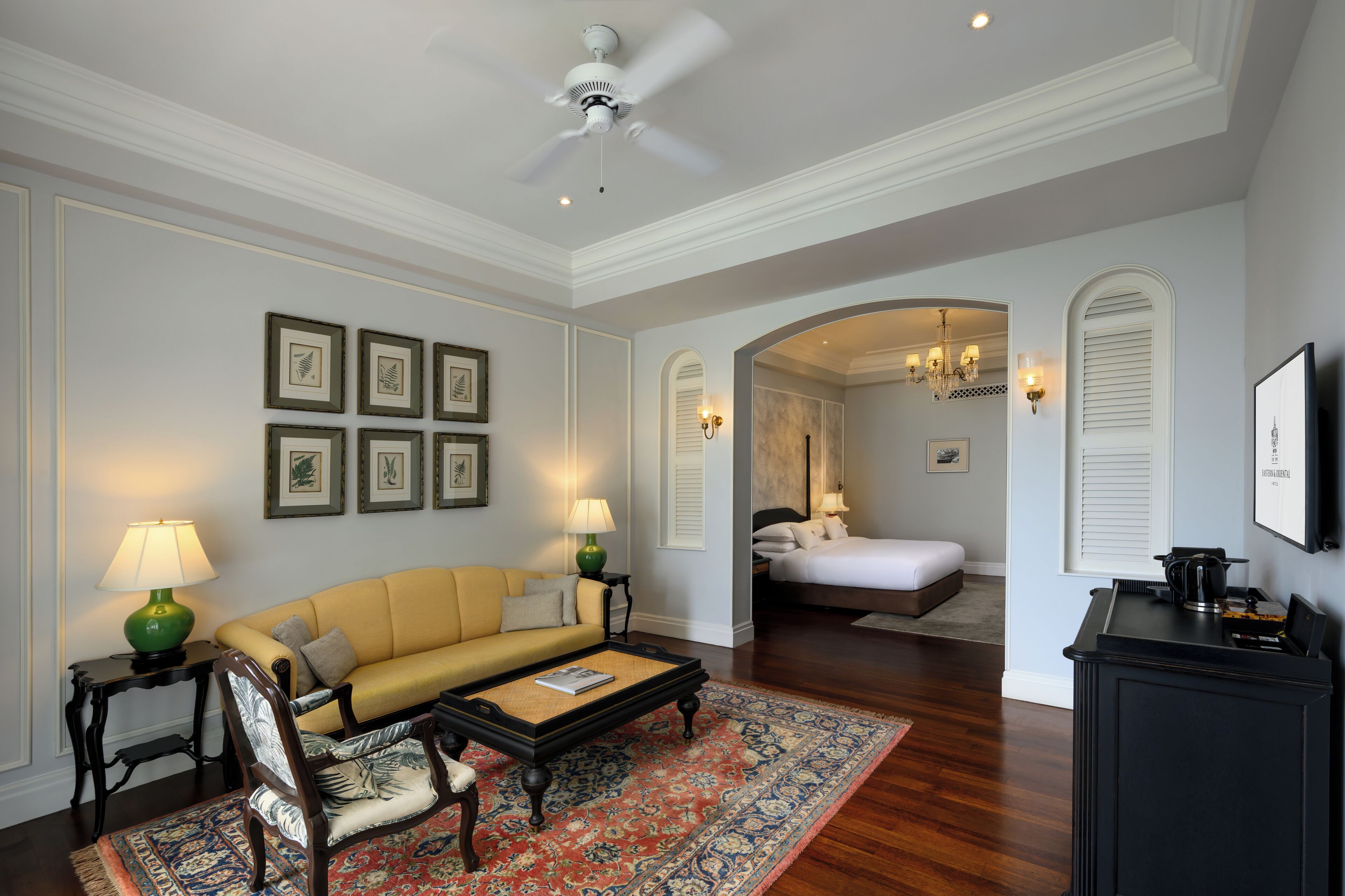 Un salón con sofá, sillones, una mesa de centro y un tapete estampado. En la habitación contigua hay una cama con una mesita de noche y una lámpara.