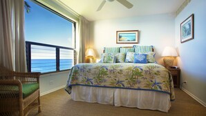 A bedroom with a large bed, two bedside lamps, a wicker chair, and a view of the ocean.