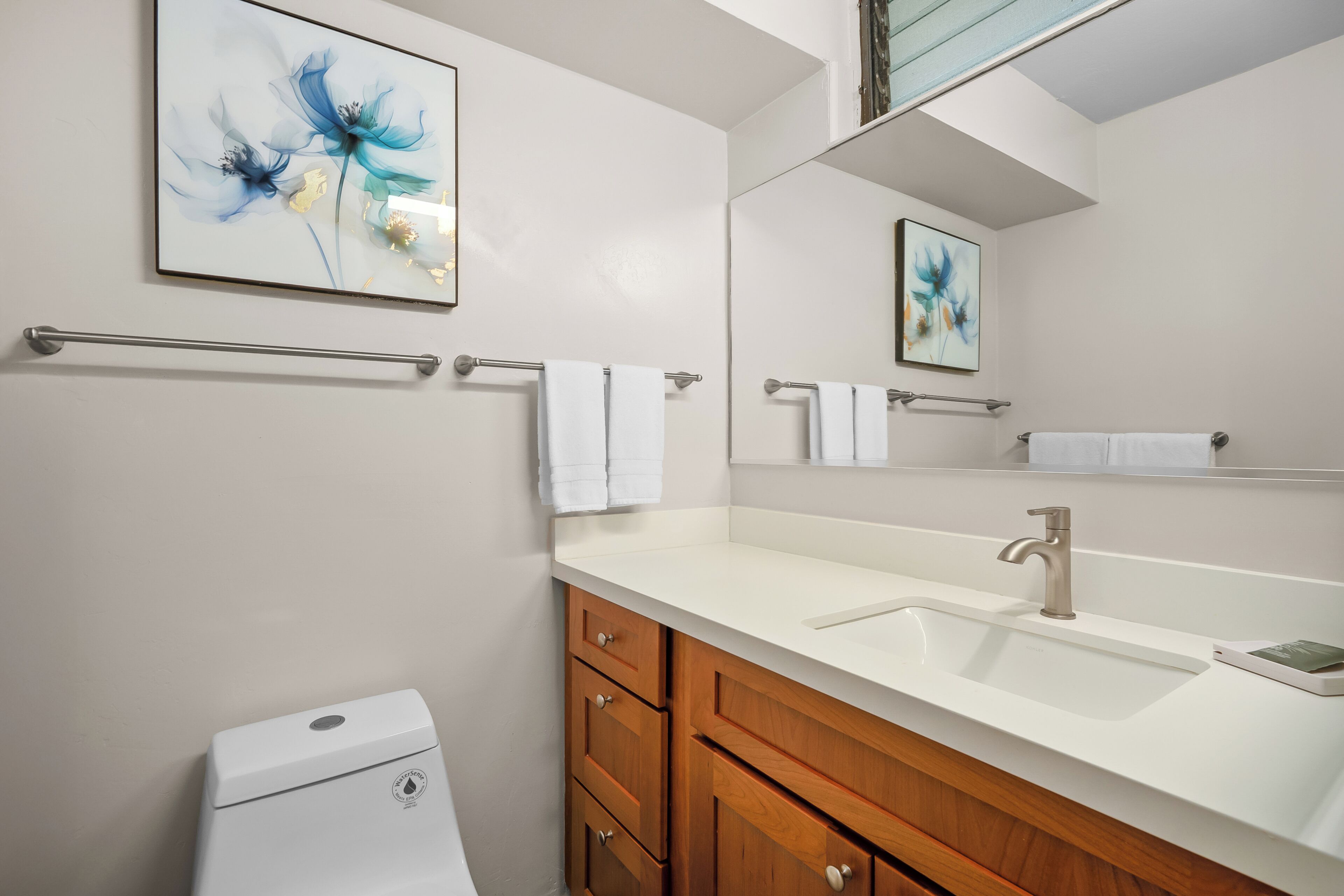 A bathroom with a white sink, wooden vanity, a toilet, and a mirror with a blue floral artwork.