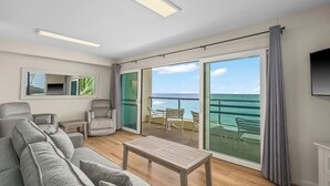 A modern living room with a sofa, chairs, a table, and a balcony with a view of the ocean.