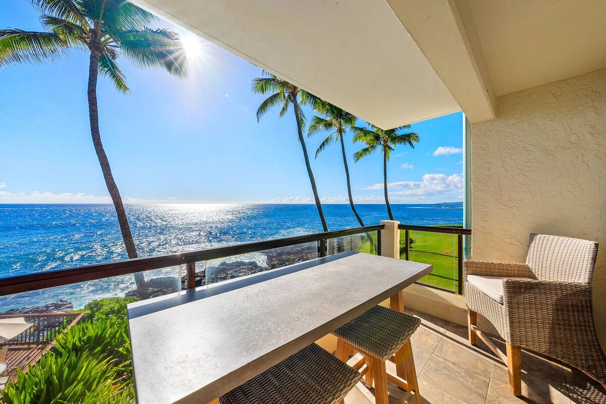 A balcony with a view of the ocean, a table, and two wicker chairs.