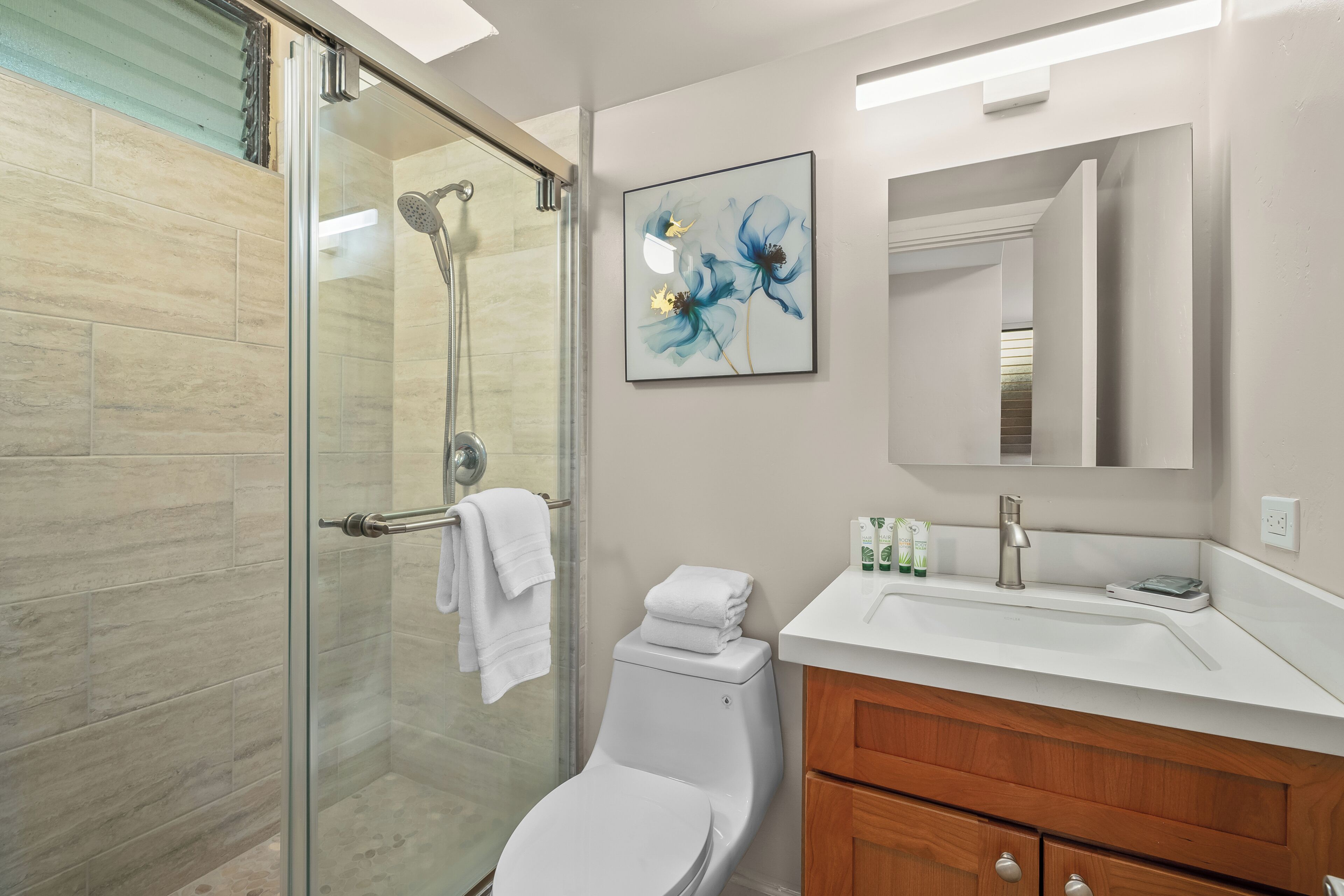 A modern bathroom with a glass shower enclosure, a white toilet, a wooden vanity with a mirror, and a framed floral artwork on the wall.