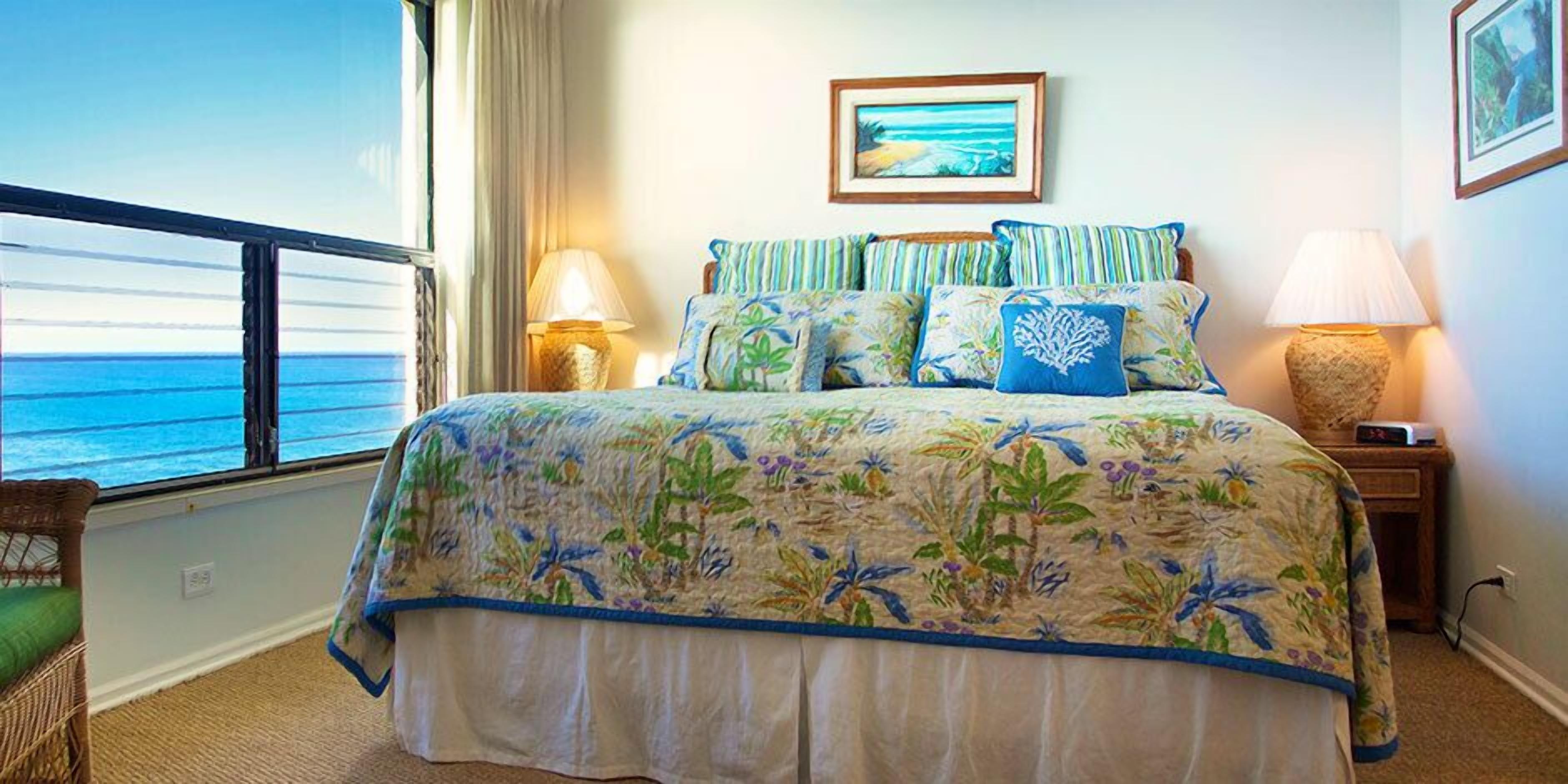 Une chambre avec une grande fenêtre, un lit recouvert d’une couette à motifs floraux, un tableau encadré au mur et une table de chevet avec une lampe.