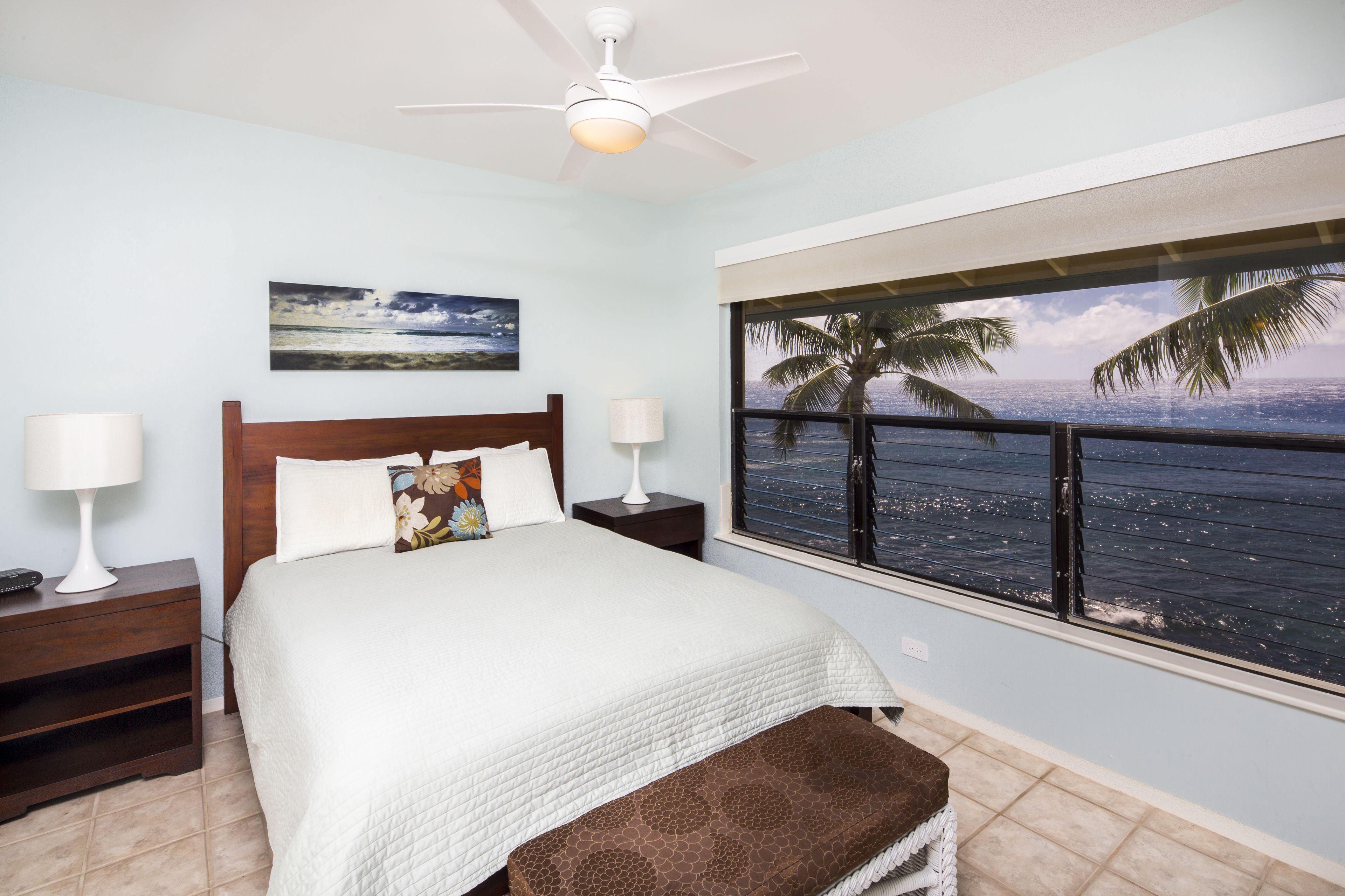 A bedroom with a bed, nightstands, a ceiling fan, a window with a view of the ocean and palm trees, and a painting on the wall.