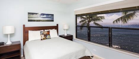A bedroom with a bed, nightstands, a ceiling fan, a window with a view of the ocean and palm trees, and a painting on the wall.