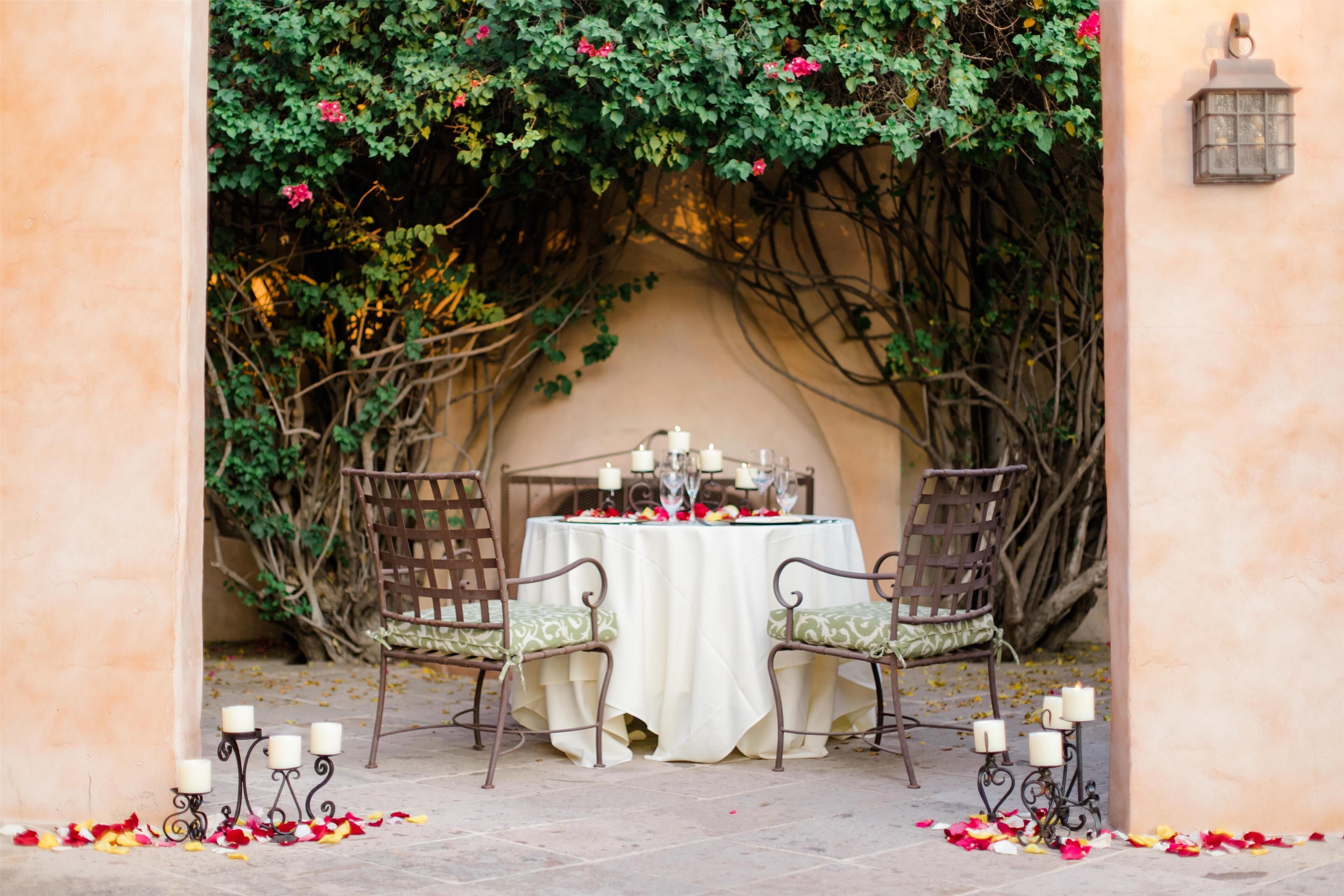 An outdoor dining area with a table set for two, surrounded by chairs, and a decorative archway with greenery.