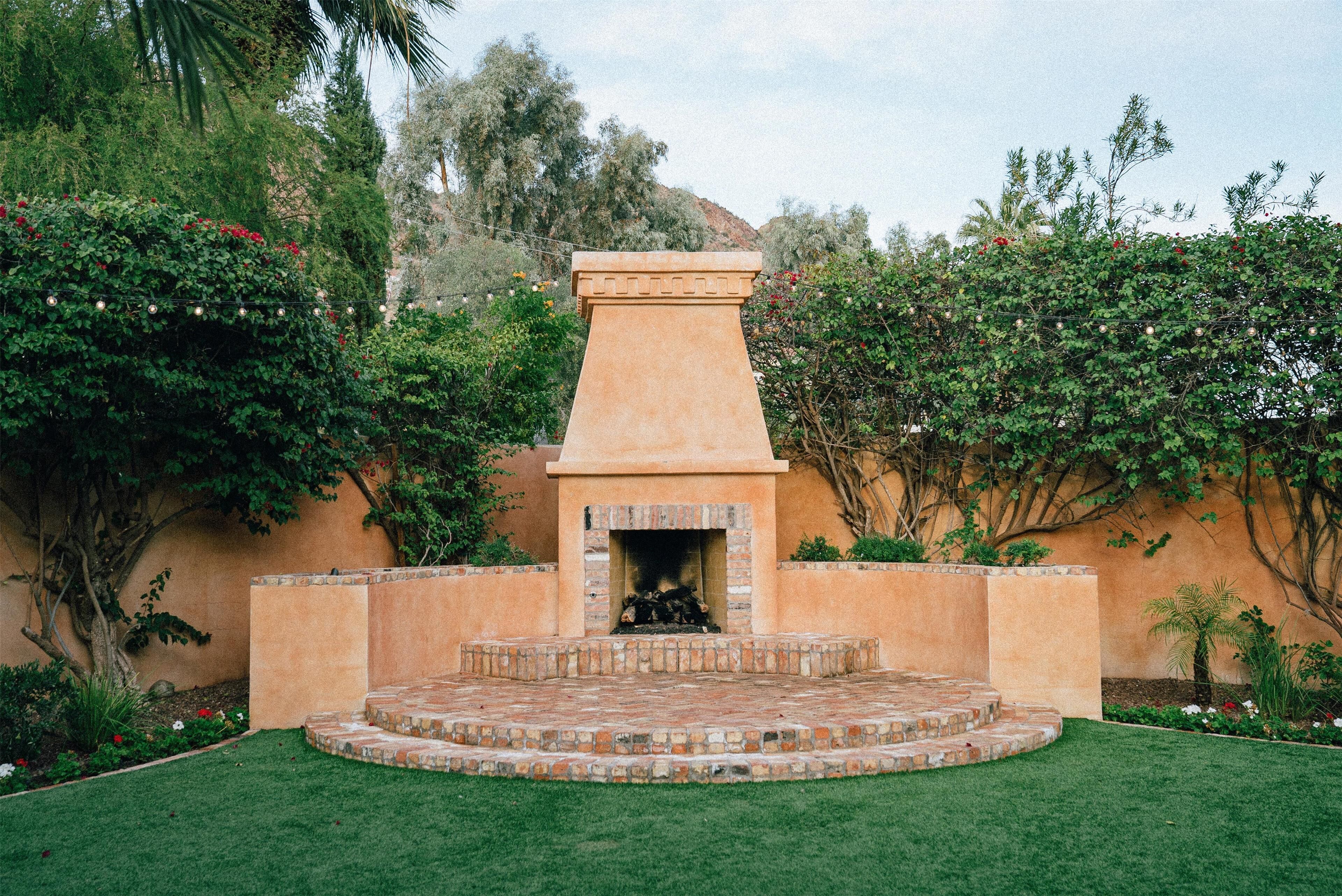 Una chimenea de piedra con detalles de ladrillo, rodeada por un jardín bien cuidado con césped y diversas plantas.