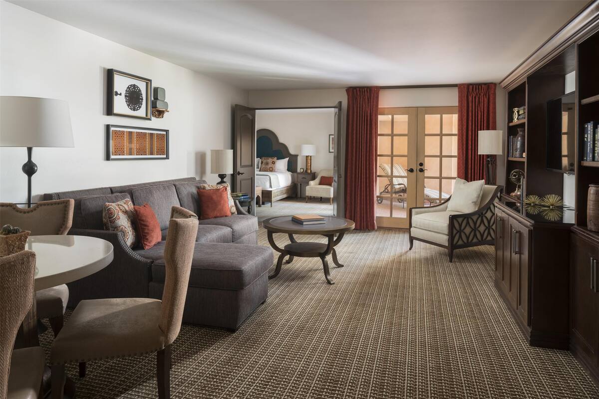 Ein geräumiges Wohnzimmer mit grauem Sofa, einem runden Couchtisch und einem Essbereich mit Stühlen. Im Hintergrund sind ein Schlafzimmer mit Bett sowie ein Badezimmer mit Dusche und Toilette zu sehen.