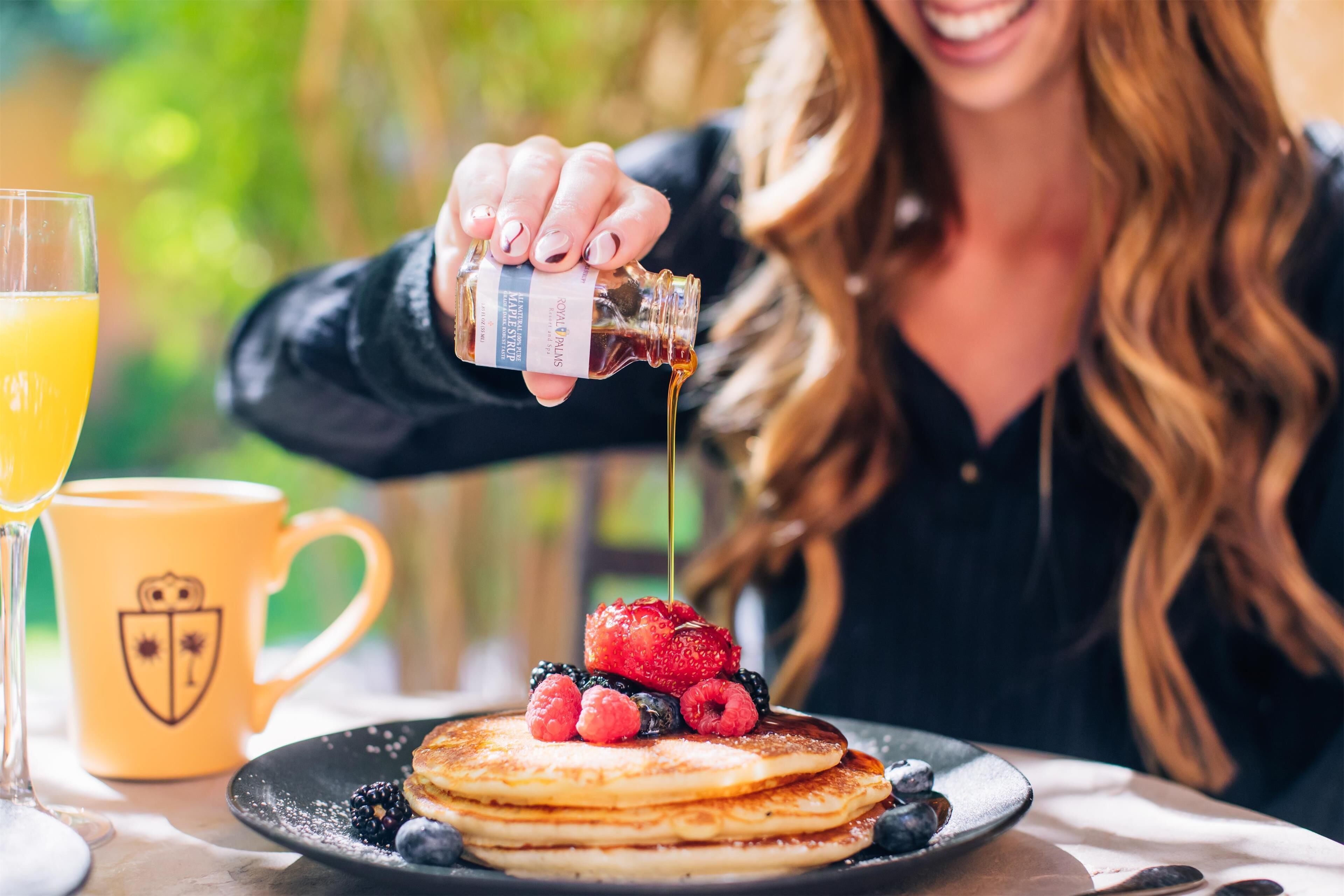 A breakfast setting with pancakes topped with berries and syrup, a yellow mug, and a glass of orange juice.