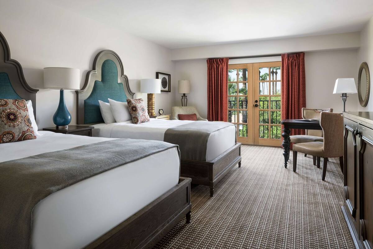 Ein Hotelzimmer mit zwei Betten, einem Schreibtisch, einem Stuhl und Blick ins Freie durch eine Glastür.