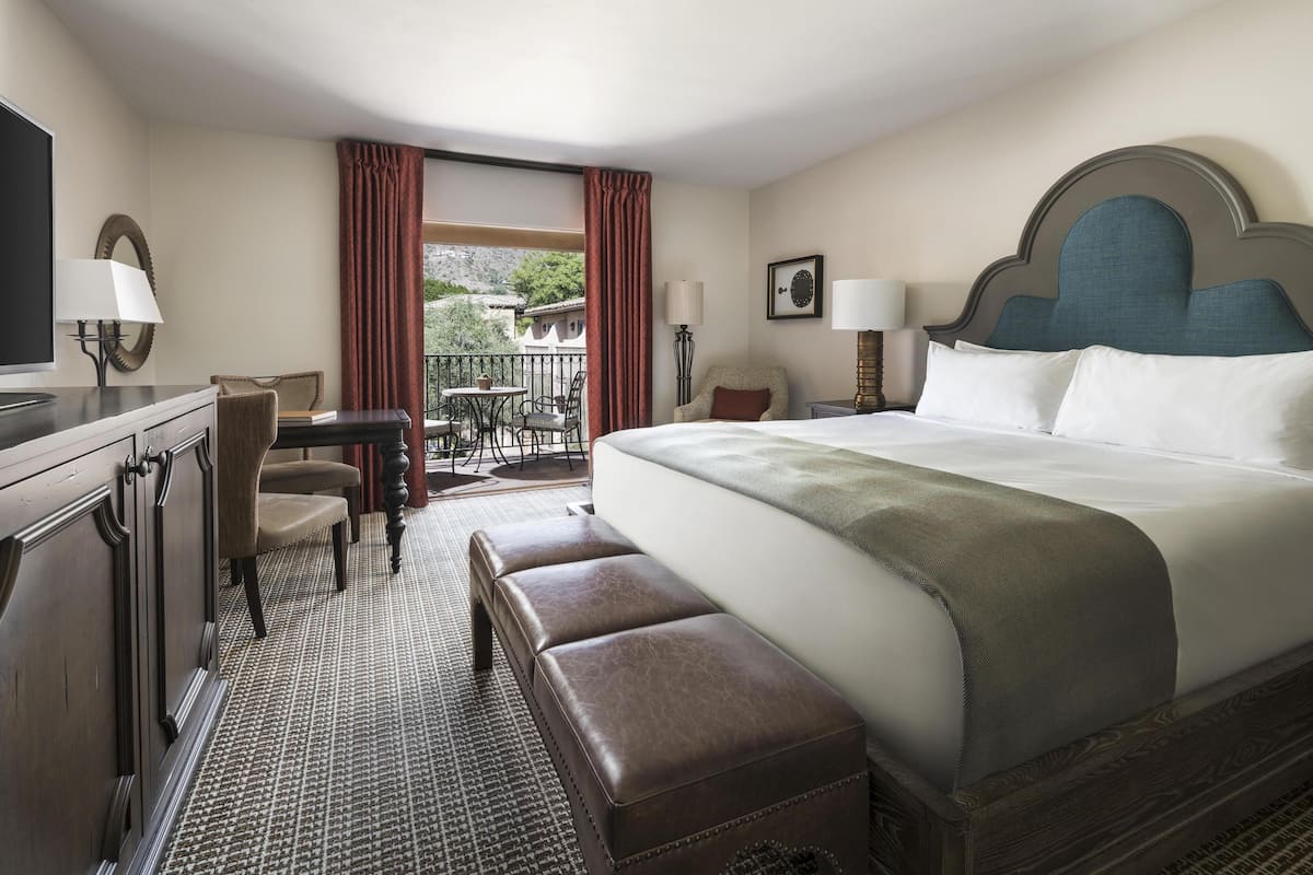 Ein Hotelzimmer mit einem großen Bett, einem Schreibtisch mit Stuhl, einem kleinen Balkon mit Tisch und Stühlen sowie einem Fernseher.