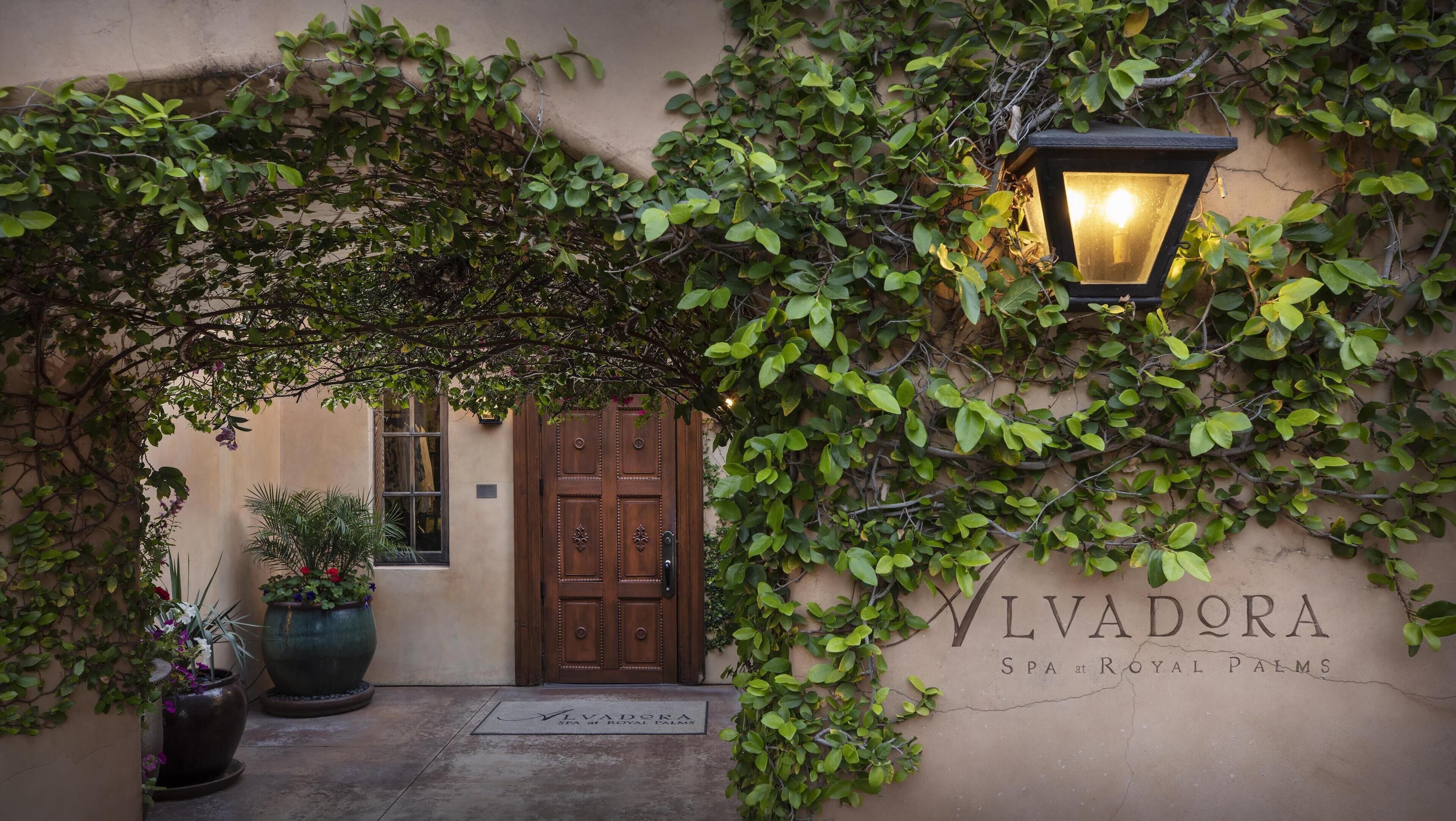 A building entrance with a wooden door, a potted plant, and a wall-mounted sign reading "VLVADORA SPA at ROYAL PALMS".