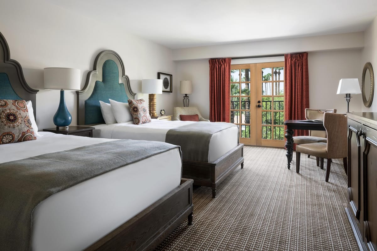 Ein Hotelzimmer mit zwei Betten, einem Schreibtisch, einem Stuhl, einem großen Fenster mit Vorhängen und Blick auf Grünfläche.