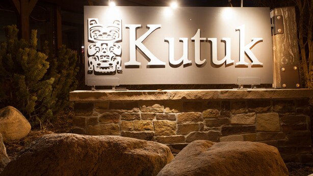 Une enseigne portant le mot « Kutuk » est illuminée la nuit, placée devant un mur de pierre et entourée de gros rochers.