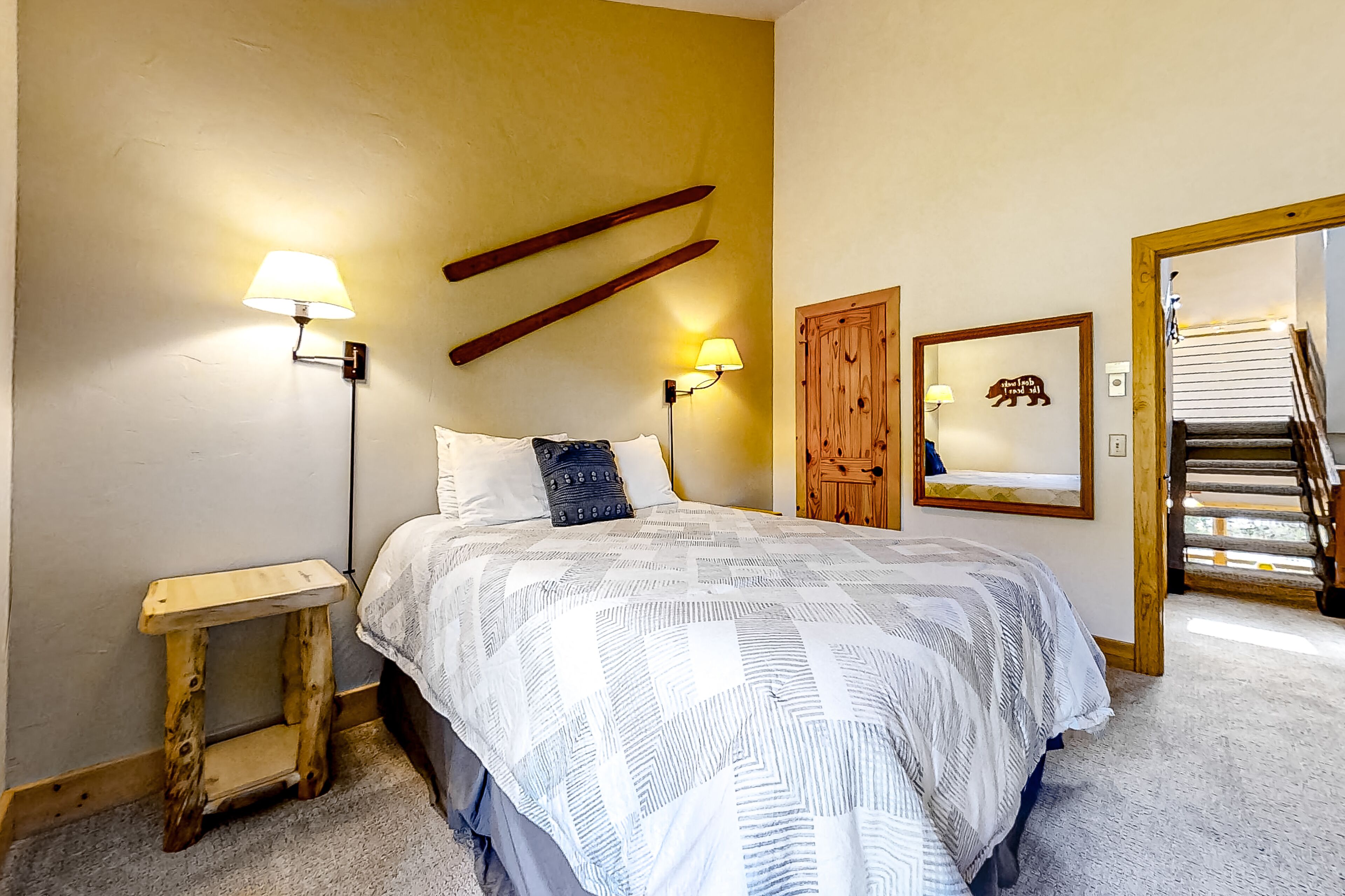 A bedroom with a bed, two bedside lamps, a wooden headboard, a small wooden nightstand, a mirror, and a staircase leading to an upper floor.