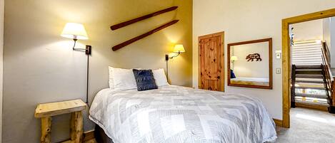 A bedroom with a bed, two bedside lamps, a wooden headboard, a small wooden nightstand, a mirror, and a staircase leading to an upper floor.