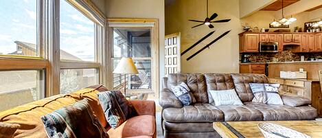 A spacious living room with a large window, a ceiling fan, and a kitchen area with wooden cabinets and a microwave.