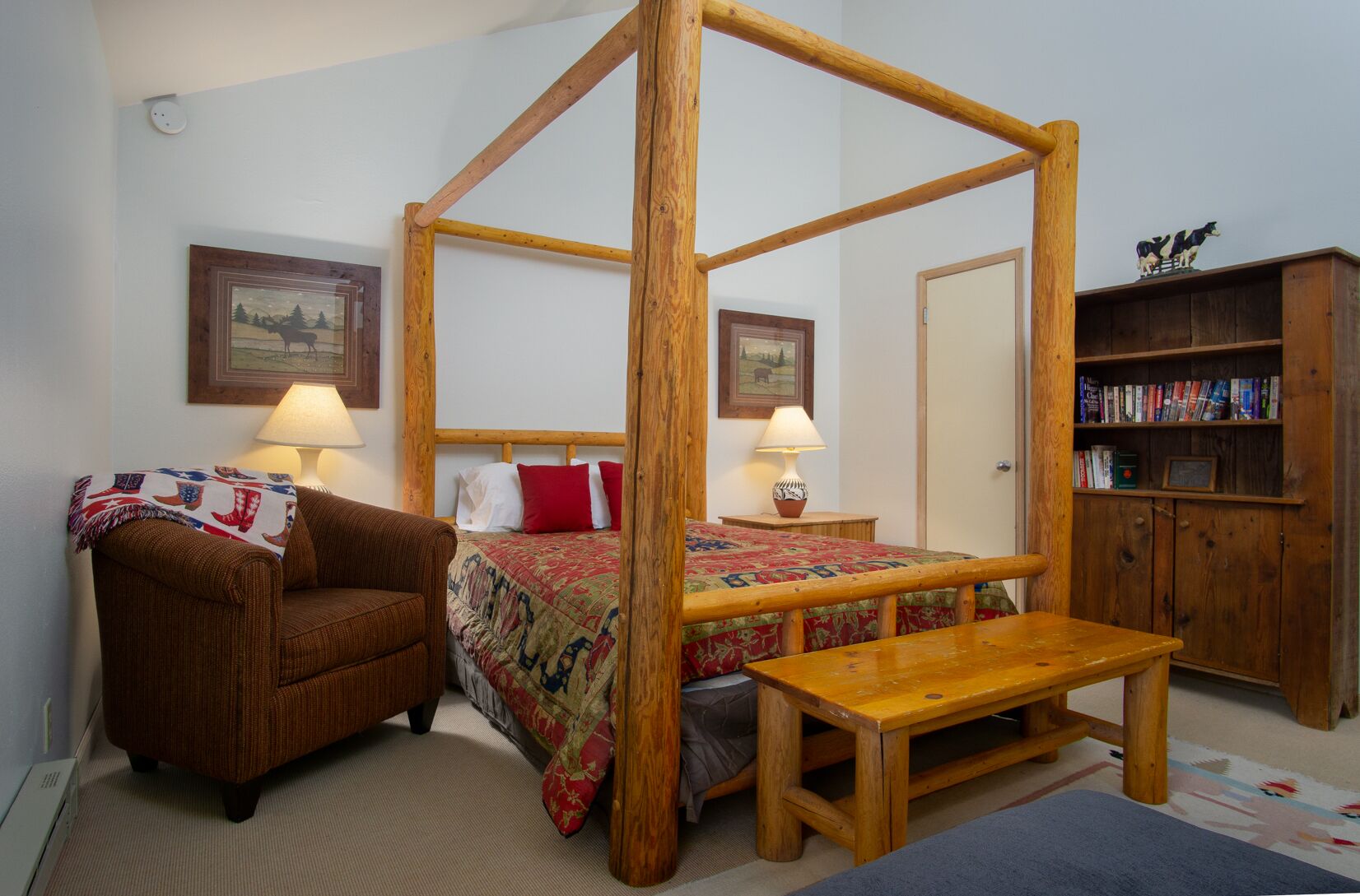Una cama con dosel y estructura de madera, un sillón marrón, una mesita de noche de madera con una lámpara, una estantería y una puerta.