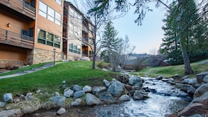 Un bâtiment moderne avec une façade en pierre, un espace verdoyant et un ruisseau bordé de rochers.