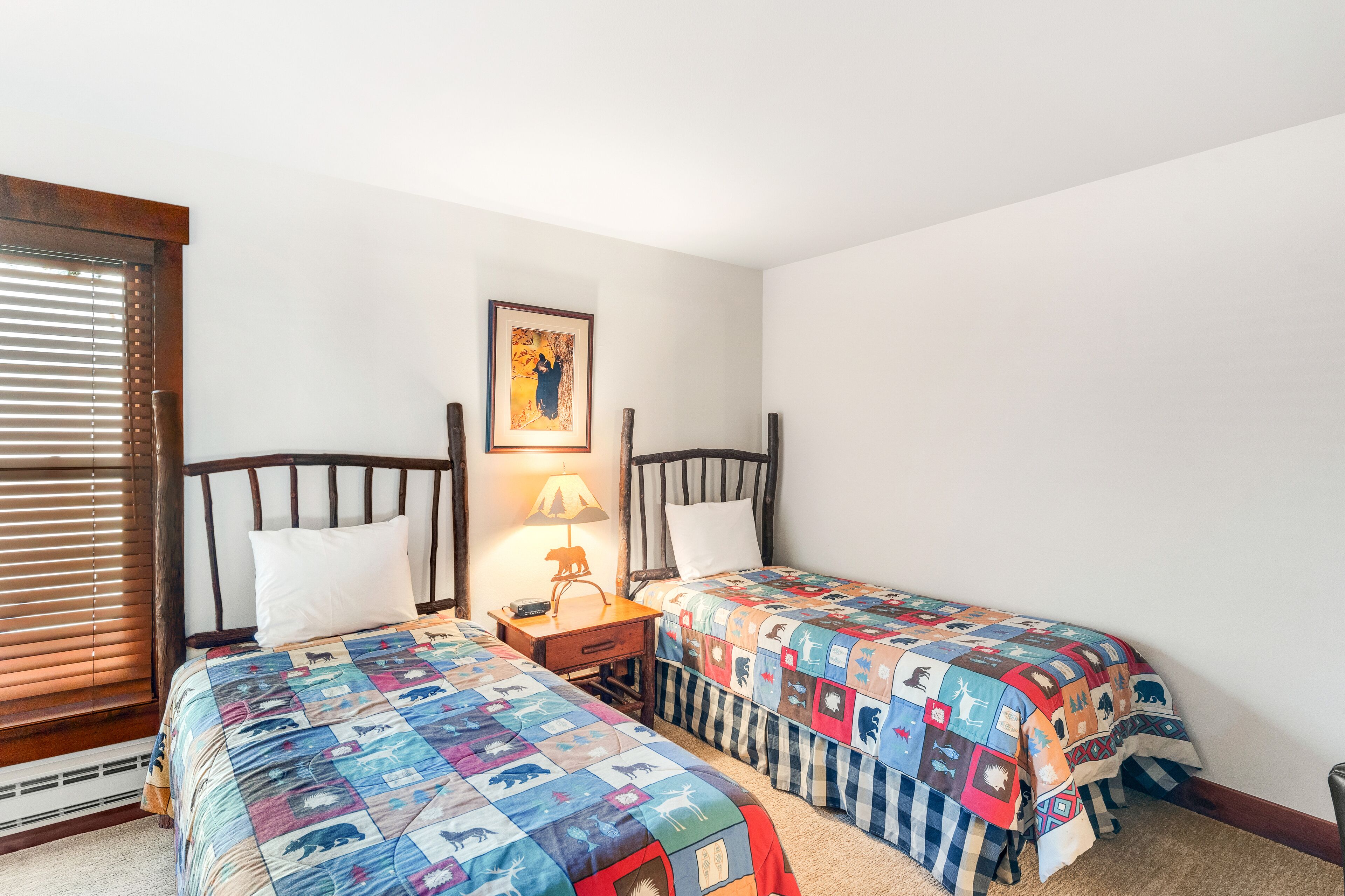 Un dormitorio con dos camas individuales, una mesita de noche con lámpara, un cuadro enmarcado en la pared y una ventana con persianas.
