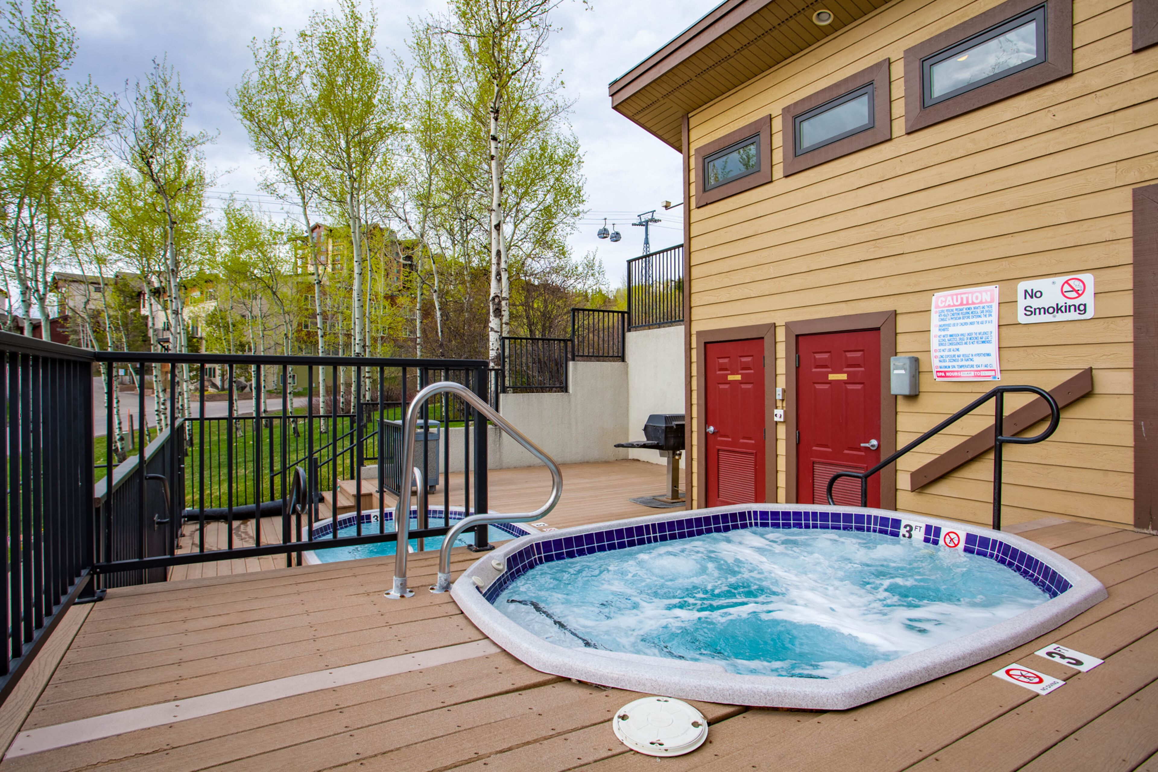 A hot tub on a wooden deck with a pool, surrounded by a fence and trees.