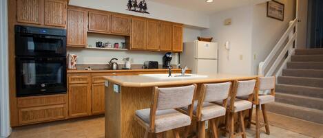 A kitchen with wooden cabinets, a built-in oven, a refrigerator, and a bar area with chairs.