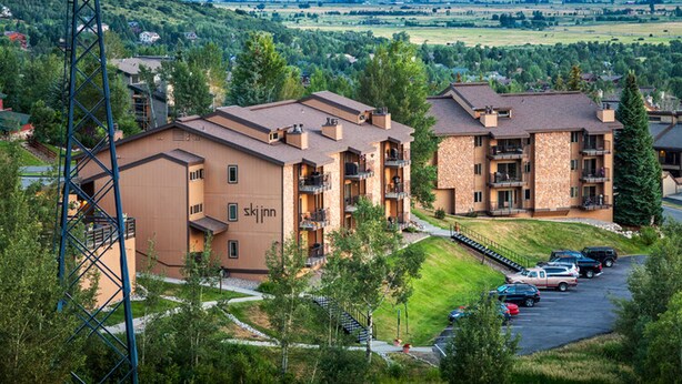 A multi-story hotel with a stone facade and balconies, surrounded by greenery and a view of distant mountains. - Ski Inn Condos (Steamboat Springs)