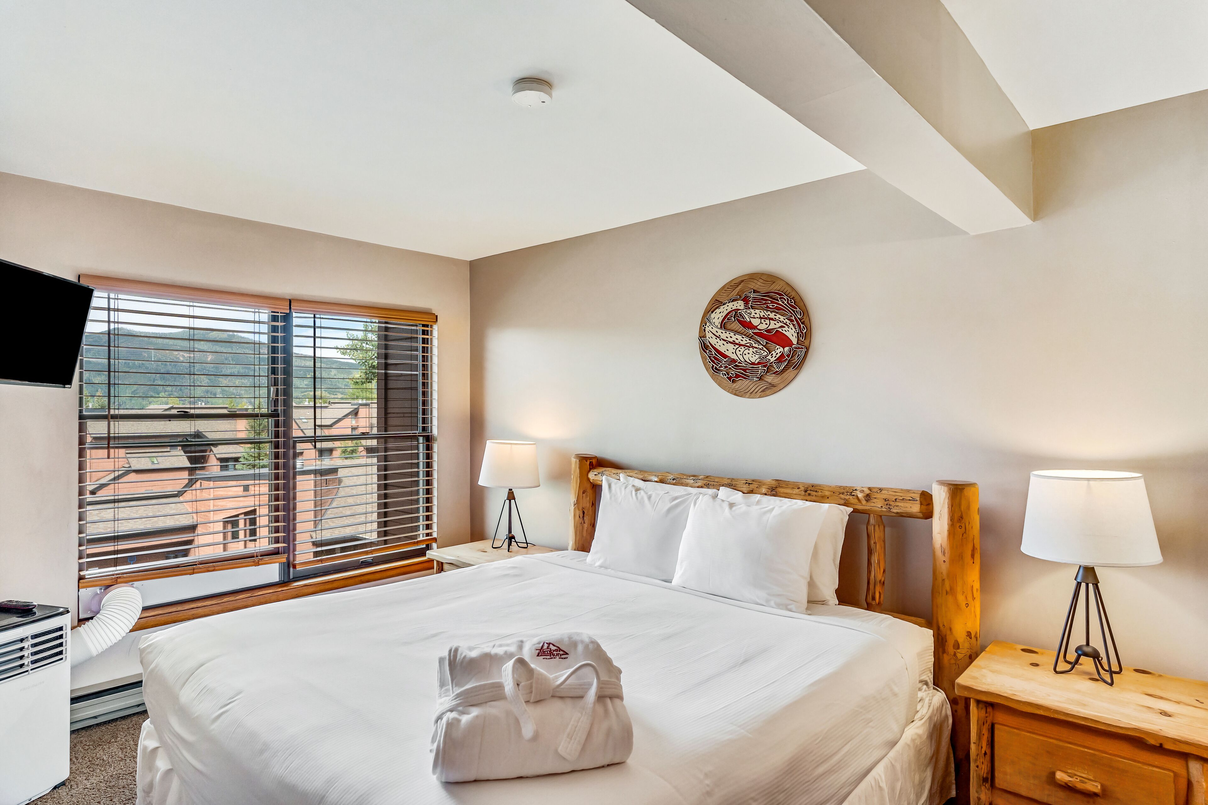 A hotel room with a large bed, wooden headboard, white bedding, a window with blinds, a wall-mounted artwork, a bedside lamp, and a wooden nightstand.
