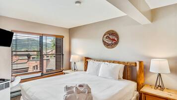 A hotel room with a large bed, wooden headboard, white bedding, a window with blinds, a wall-mounted artwork, a bedside lamp, and a wooden nightstand.