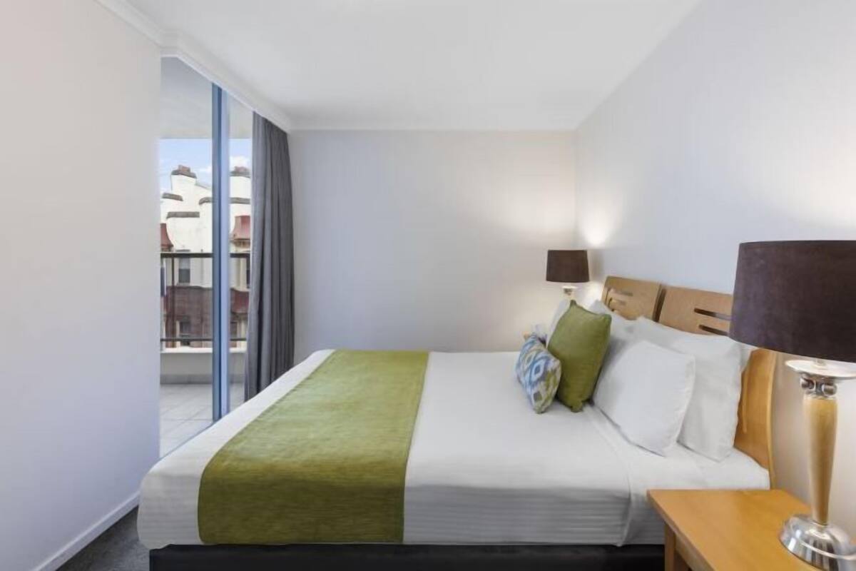 A hotel room with a bed, a bedside table with a lamp, a window with curtains, and a view of buildings outside.