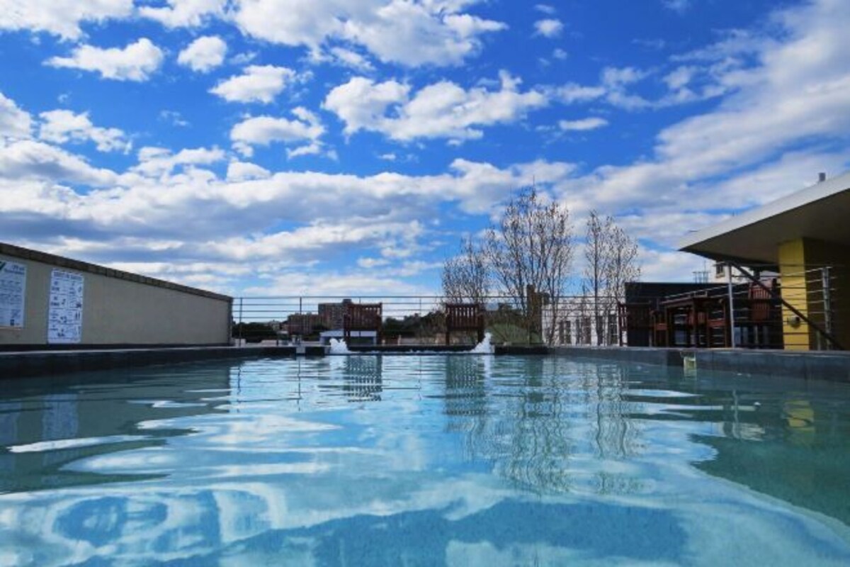 Une piscine à l’eau claire, entourée de bâtiments et surplombée d’un ciel partiellement nuageux.