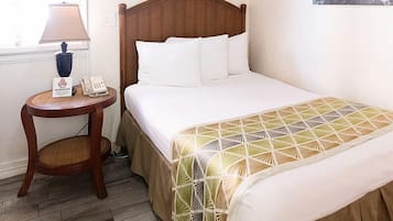 Ein ordentlich bezogenes Bett mit weißen Leinentüchern und einem Bettbezug mit geometrischem Muster, ein Nachttisch mit einer Lampe und einem Telefon sowie ein Wandbild.