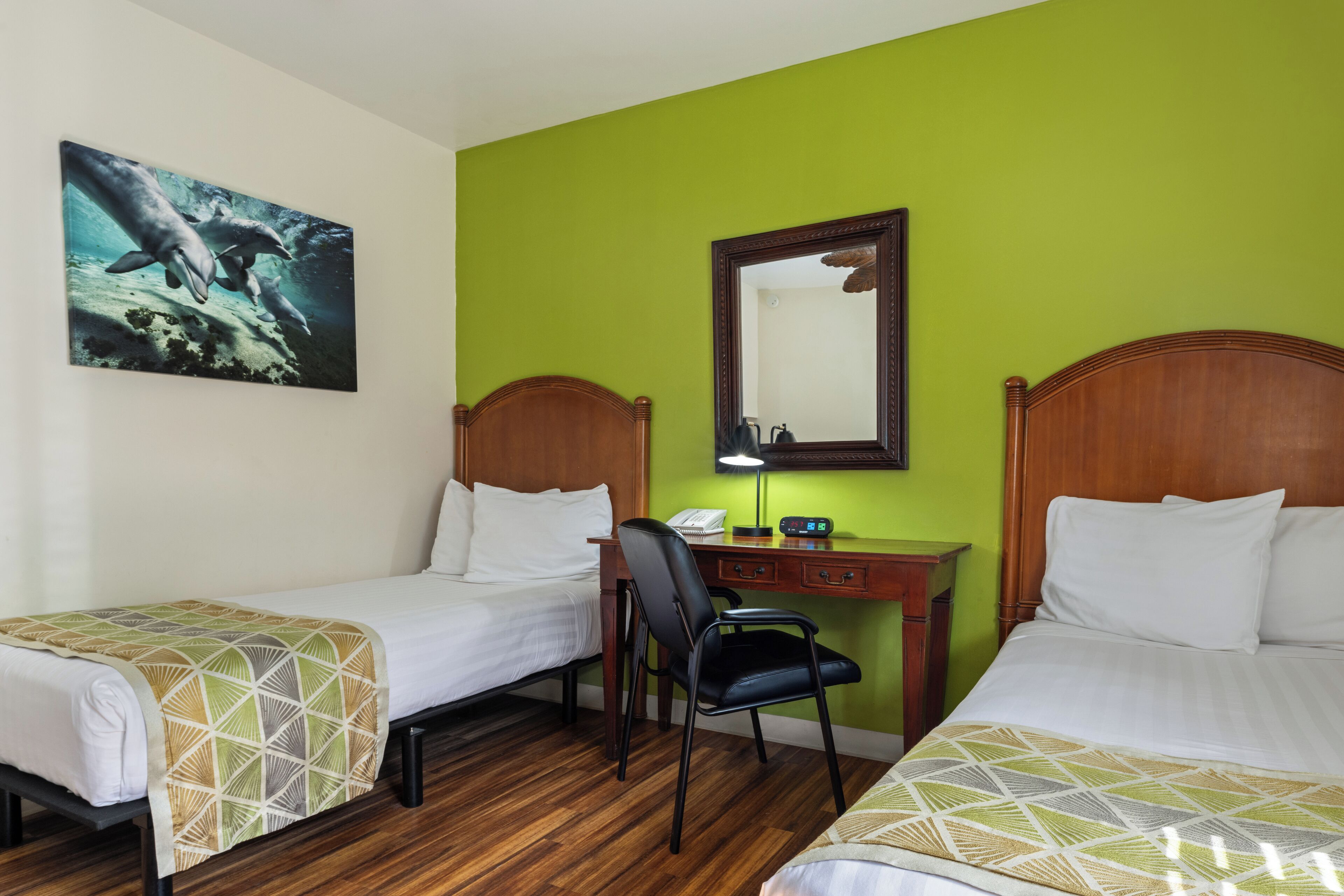 A hotel room with two single beds, a desk with a chair, a mirror, and a painting of dolphins on the wall.