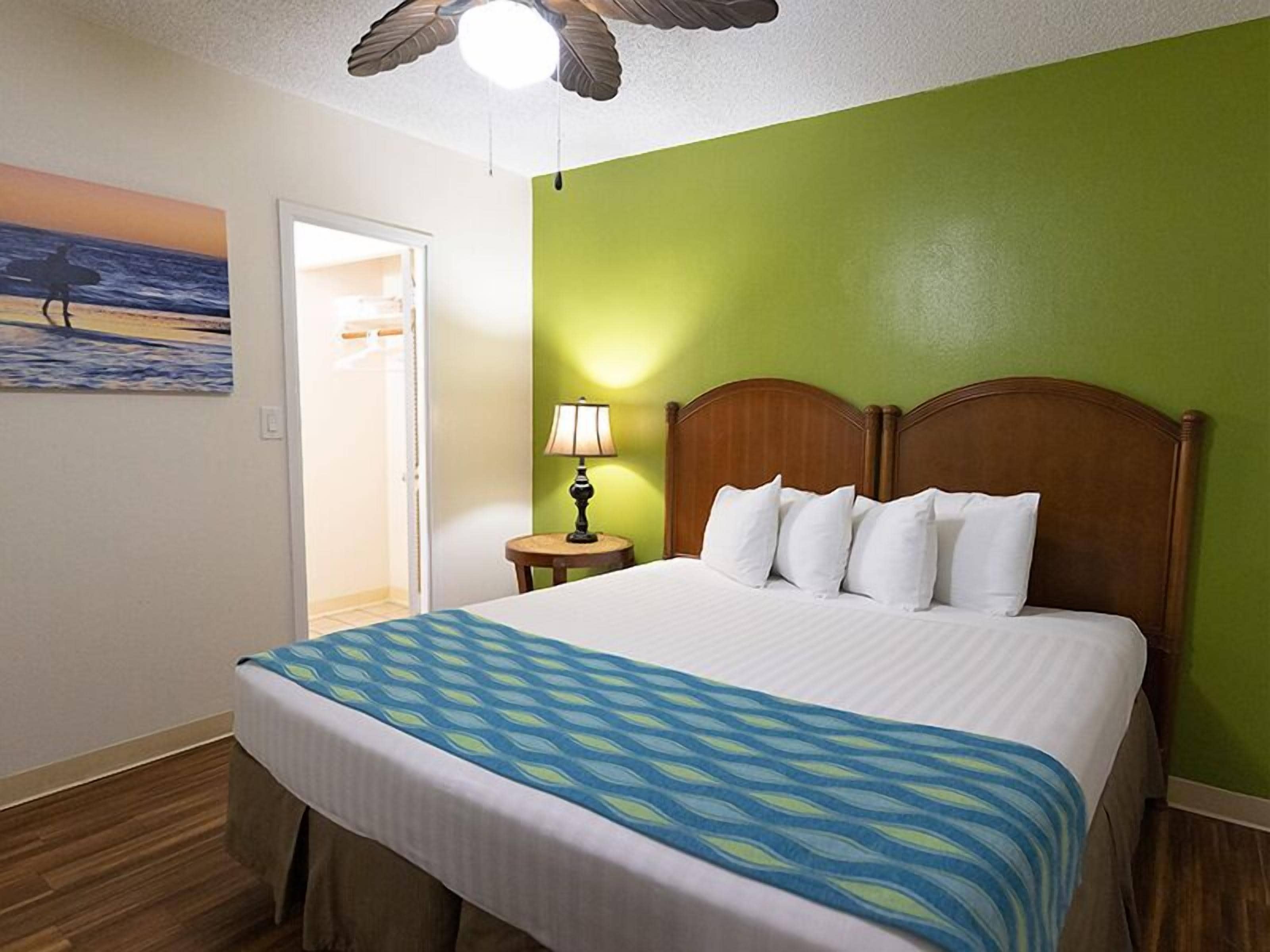 Une chambre d’hôtel avec un lit, une table de chevet avec une lampe, un tableau représentant une scène de plage et un ventilateur de plafond.