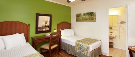 A hotel room with two beds, a desk, a chair, a ceiling fan, a painting, and a bathroom visible through an open door.