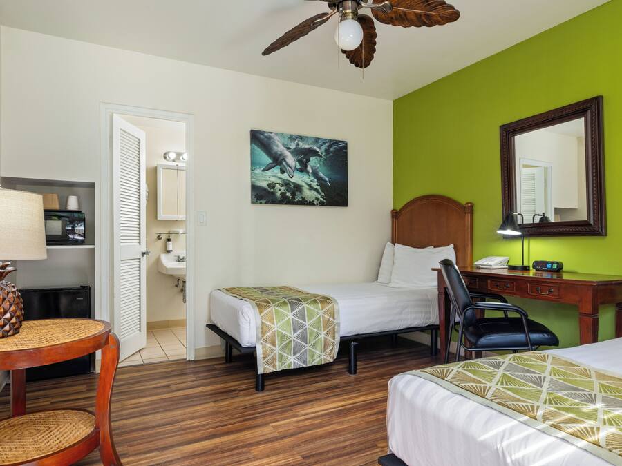 A hotel room with two beds, a desk, a chair, a ceiling fan, a painting, and a bathroom visible through an open door.