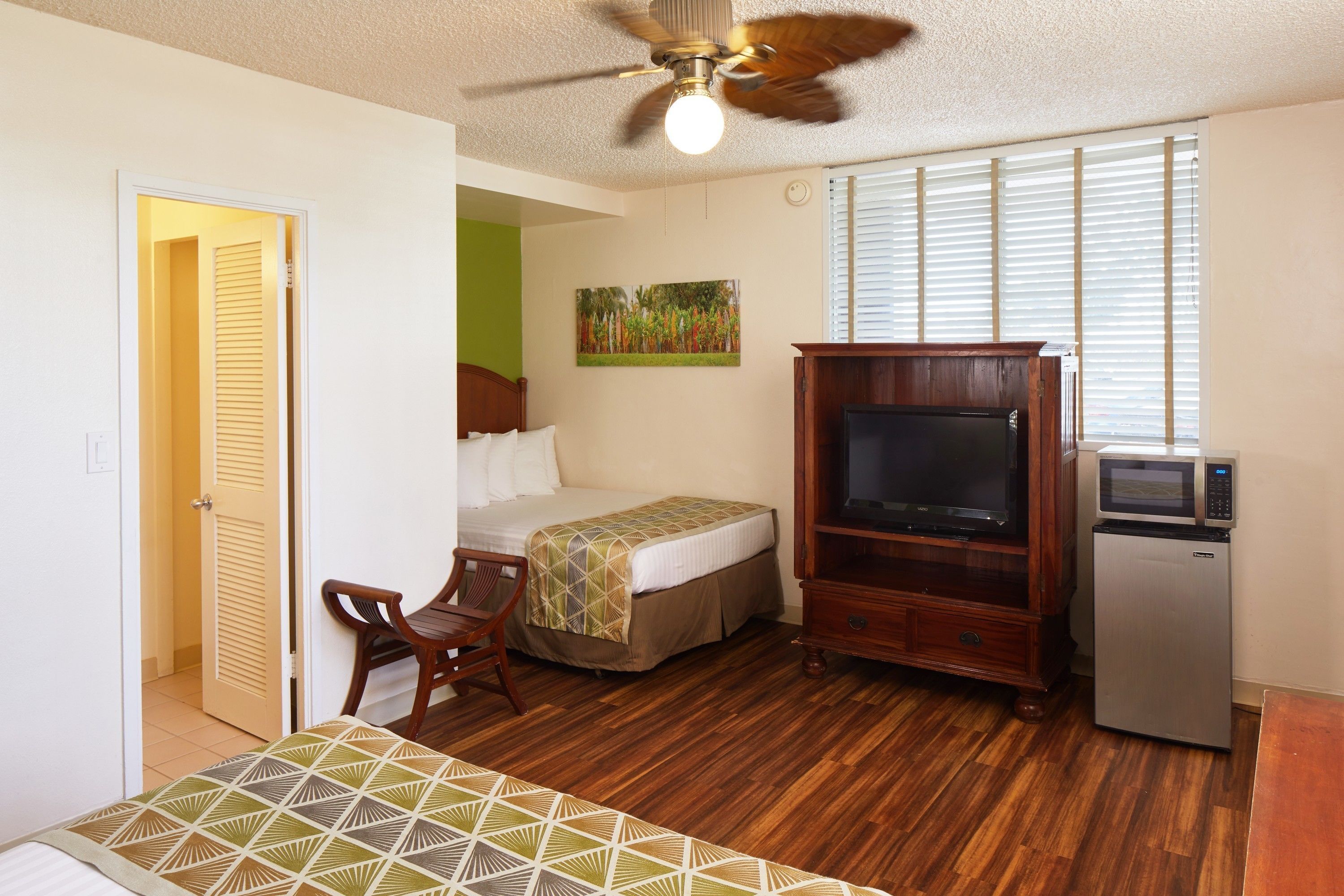 Habitación de hotel con cama, escritorio, televisor, refrigerador y ventilador de techo.