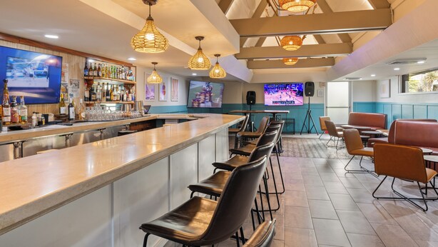 A bar area with a long counter, multiple bar stools, and overhead wicker lamps. There are televisions mounted on the walls and a speaker near the bar.