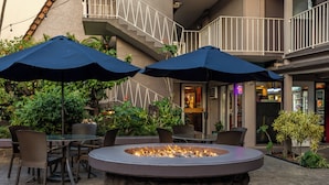 An outdoor patio area with a circular stone fire pit, surrounded by chairs and tables, and two large umbrellas providing shade.